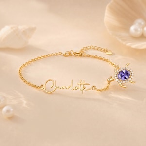 May include: A gold-tone chain bracelet featuring the name Charlotte in cursive script. The bracelet includes a turtle-shaped charm with a central oval purple gemstone surrounded by small clear stones, connected to a delicate link chain.