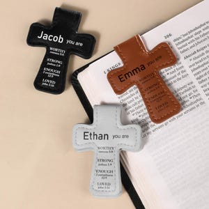 May include: Three cross-shaped bookmarks in black, brown, and white. Each bookmark has a name printed on it, along with the words "Worthy," "Strong," "Enough," and "Loved," and corresponding Bible verses. The bookmarks are made of a leather-like material.