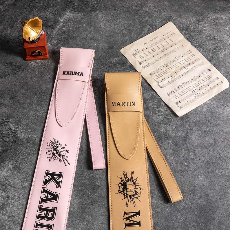 May include: Two guitar straps, one pink with the name "KARIMA" and the other brown with the name "MARTIN". Both straps have a graphic design of a fist holding a drumstick.