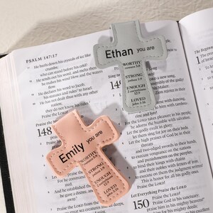 May include: Two cross-shaped leather bookmarks. One is light gray with "Ethan you are" and inspirational text. The other is peach with "Emily you are" and similar text. Both are on an open book.