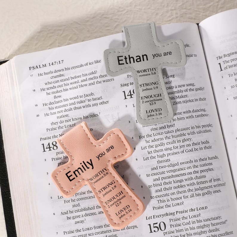 May include: Two cross-shaped leather bookmarks. One is light gray with "Ethan you are" and inspirational text. The other is peach with "Emily you are" and similar text. Both are on an open book.