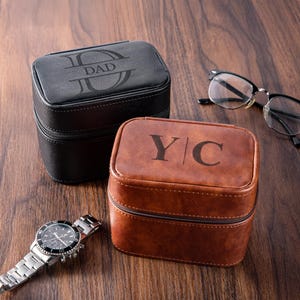 Puede incluir: Dos estuches rectangulares para relojes, uno negro y otro marrón, con cierres de cremallera. El estuche negro está grabado con "DAD", y el marrón tiene "YC". Un reloj de pulsera plateado y gafas también están presentes, todo sobre una superficie de madera.