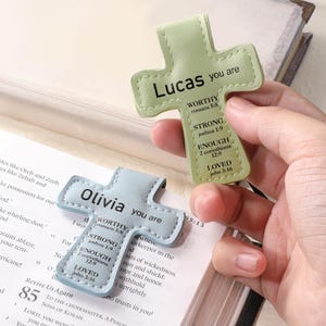 May include: Two leather cross-shaped bookmarks, one light green and one light blue, are displayed open on a book. The green bookmark says "Lucas you are" with inspirational text. The blue bookmark says "Olivia you are" with similar text.