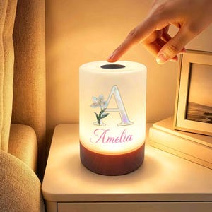 May include: A cylindrical bedside lamp with a warm glowing white shade featuring a large letter A, a small floral illustration, and the name Amelia printed in pink. The lamp has a dark wood-toned base and a circular touch-sensitive top.