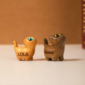 Könnte beinhalten: Zwei Holzkatzenfiguren, eine hellbraun mit dem eingravierten Namen "LOLA" und die andere dunkelbraun mit dem eingravierten Namen "Penny".