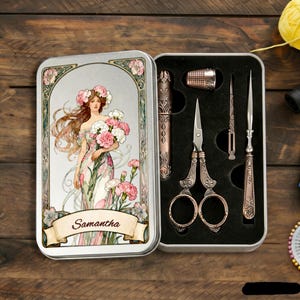 Pode incluir: Um estojo de costura retangular de metal com a ilustração de uma mulher segurando flores e o nome Samantha impresso numa faixa. O estojo aberto contém um dedal, tesoura, um estojo de agulhas e um furador num encaixe de espuma preta.
