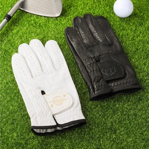 May include: A pair of golf gloves, one white and one black, are displayed on a green surface. The white glove has a gold emblem and black trim. The black glove has a textured palm and the initials "FT" on the wrist. A golf ball and club are also visible.