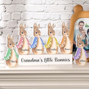 Könnte beinhalten: Ein Set aus fünf Holz-Hasenfiguren mit verschiedenen farbigen Jacken, jede mit einem Namen darunter: Oliver, Sophia, James, Emilia und Mason. Die Hasen sind in einer Reihe auf einer weißen Holzbasis mit dem Text "Grandma's Little Bunnies" angeordnet.