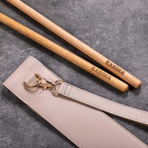 May include: Two wooden chopsticks with the word "KARIMA" engraved on them, lying on a light beige leather strap with a gold clasp.