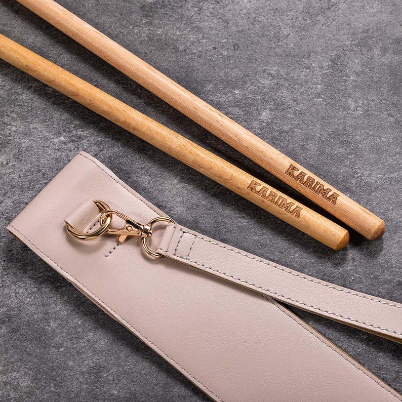 May include: Two wooden chopsticks with the word "KARIMA" engraved on them, lying on a light beige leather strap with a gold clasp.