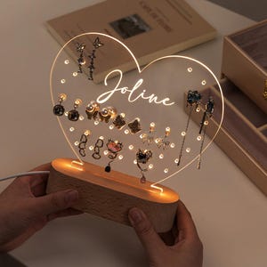 Pode incluir: Um suporte para brincos de acrílico em forma de coração com uma base de madeira. O suporte tem uma luz no interior e o nome "Joline" está escrito na parte frontal. O suporte está cheio de vários brincos.