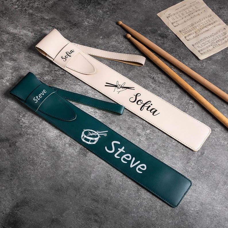 May include: Two personalized drum stick holders, one in green with a drum and sticks design and the text "Steve", and one in beige with the text "Sofia".
