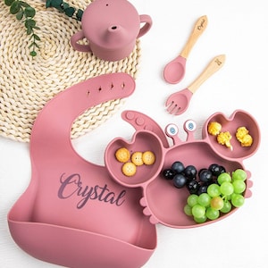 May include: A pink silicone crab-shaped plate with three compartments, a pink silicone bib with the name "Crystal" printed on it, and two wooden spoons with pink silicone tips.
