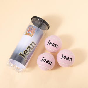 May include: Three pink tennis balls with the name "Jean" printed on each ball. The balls are next to a clear plastic container with a black lid. The container has a label with the name "Jean" printed on it.