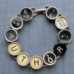 Könnte beinhalten: Ein silbernes Armband aus Schreibmaschinentasten. Die Tasten buchstabieren das Wort "AUTHOR" und beinhalten weitere Tasten mit den Wörtern "MARGIN RELEASE" und den Symbolen "@".