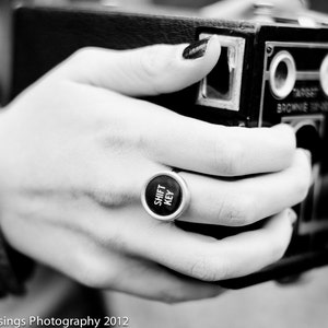 May include: A black and silver vintage camera with a ring on the hand holding it. The ring is silver and has the words "Shift Key" on it.
