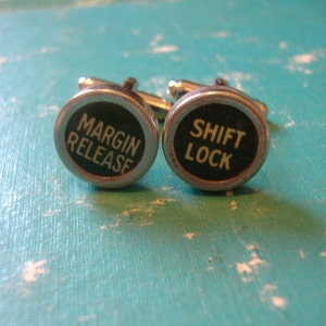 May include: A pair of cufflinks with black round buttons. The left cufflink has the text "MARGIN RELEASE" in white lettering. The right cufflink has the text "SHIFT LOCK" in white lettering.
