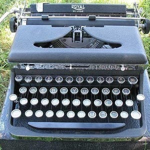 May include: A black Royal De Luxe typewriter with a standard keyboard layout. The typewriter is sitting on a black case and is surrounded by green grass.