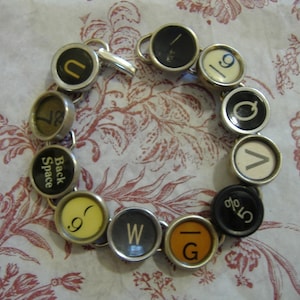 Könnte beinhalten: Ein Armband aus Schreibmaschinentasten. Die Tasten sind durch silberne Metallglieder verbunden. Die Tasten haben schwarze und weiße Beschriftungen und beinhalten Buchstaben, Zahlen und Symbole. Das Armband befindet sich auf einem weißen Hintergrund mit einem Blumenmuster.