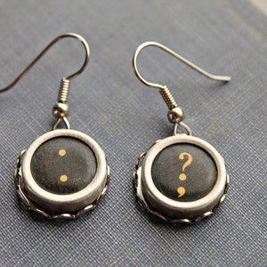 May include: A pair of silver earrings with black typewriter keys. The keys have gold punctuation marks: a semicolon and a question mark.