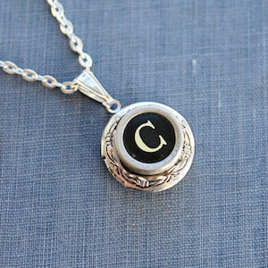 May include: Silver chain necklace with a locket pendant featuring a vintage typewriter key with the letter 'C' in white on a black background.