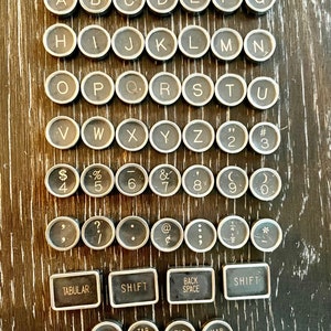 May include: A set of vintage typewriter keys with black lettering on a silver background. The keys include letters, numbers, symbols, and function keys such as Tab, Shift, Back Space, Lock, Clear, Set, and Rel.