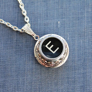 May include: Silver necklace with a locket featuring a black typewriter key with a white letter E.