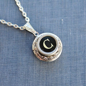 May include: Silver chain necklace with a locket pendant featuring a black typewriter key with a white letter 'C'.