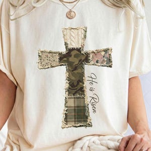 May include: Cream-colored t-shirt featuring a patchwork cross design with various patterns, including camouflage and plaid. The text "He is Risen" is printed on the right side of the cross. The shirt has short sleeves.