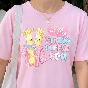 May include: A pale pink t-shirt with a cartoon design of two string cheese characters in pink robes and a blue bow. The text "in My STRING CHEESE era" is printed on the shirt in various colours.