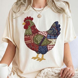 Könnte beinhalten: Cremefarbenes T-Shirt mit einem bunten Patchwork-Hühnerdesign. Das Huhn hat einen roten Kamm, gelbe Füße und einen Körper aus verschiedenen gemusterten Stoffflicken in Rot, Grün und Blau. Das Shirt hat kurze Ärmel.