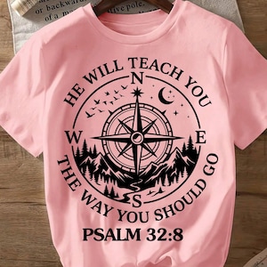 May include: A light pink t-shirt with a black compass design. The compass is surrounded by the text "He Will Teach You The Way You Should Go" and "Psalm 32:8". The design includes mountains, birds, and a crescent moon.