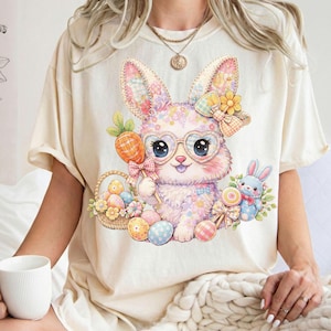 May include: Cream-colored t-shirt featuring a colorful Easter bunny design. The bunny wears heart-shaped glasses and holds a carrot lollipop. A basket of Easter eggs and a small blue bunny are also depicted.