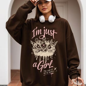 以下が含まれることがあります： ピンク色の「I'm just a Girl」の文字と王冠のグラフィックがプリントされた茶色のスウェットシャツ。 スウェットシャツは長袖とクルーネックです。 「I've had it up to here!」の文字もプリントされています。