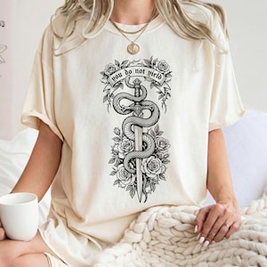 May include: Cream-colored t-shirt featuring a black and white graphic of a sword entwined by a snake, surrounded by roses. The banner above the sword reads "YOU DO NOT YIELD".