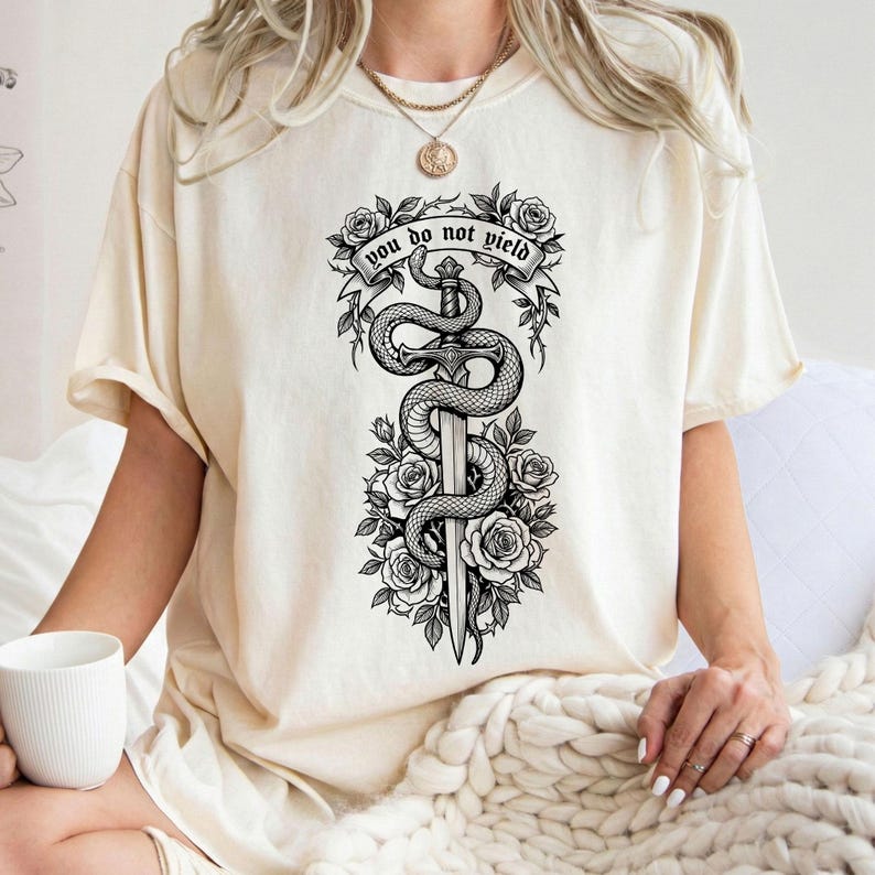 May include: Cream-colored t-shirt featuring a black and white graphic of a sword entwined by a snake, surrounded by roses. The banner above the sword reads "YOU DO NOT YIELD".