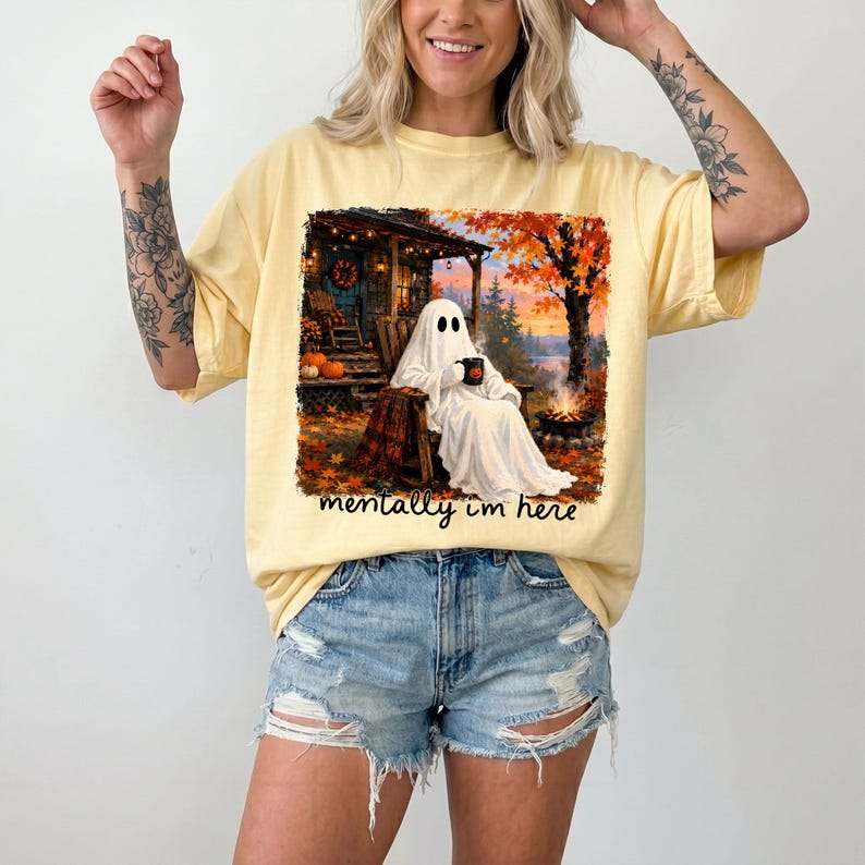 May include: A pale yellow short-sleeve t-shirt featuring a graphic print of a ghost sitting in a rocking chair on a porch, holding a mug. The scene includes autumn trees, a campfire, and pumpkins. The text "mentally im here" is printed below.