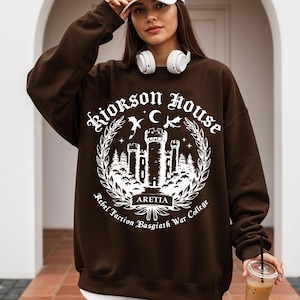Puede incluir: Una sudadera marr&oacute;n con un gr&aacute;fico blanco que presenta un castillo, una luna, estrellas y el texto "Riorkson House Rebel Faction Basgiath War College ARETIA". La sudadera tiene mangas largas y cuello redondo.