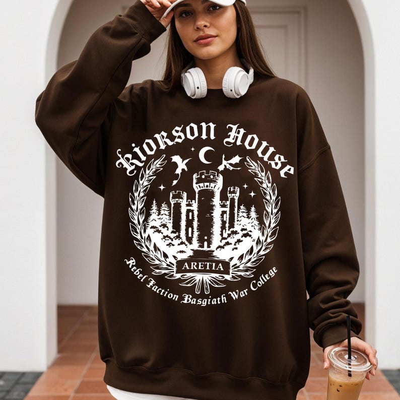 Puede incluir: Una sudadera marr&oacute;n con un gr&aacute;fico blanco que presenta un castillo, una luna, estrellas y el texto "Riorkson House Rebel Faction Basgiath War College ARETIA". La sudadera tiene mangas largas y cuello redondo.