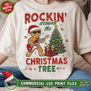 Puede incluir: Camiseta color crema con un hombre de jengibre con gafas de sol y un gorro de Papá Noel, tocando una guitarra roja. El texto dice "ROCKIN' around the CHRISTMAS TREE" en rojo y verde. También se muestra un árbol de Navidad decorado y una caja de regalo.
