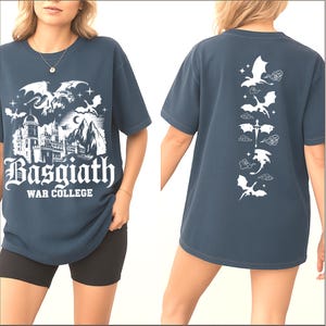 May include: A navy blue t-shirt featuring a white graphic of a castle, dragons, and the text "Basgiath War College" on the front. The back of the shirt displays a vertical line of dragon silhouettes.