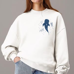 May include: White sweatshirt with a crew neck featuring a blue whale shark illustration and other sea creatures. The design includes a turtle, jellyfish, and stars. The sweatshirt is a casual, comfortable style.