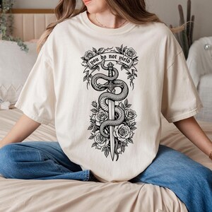 May include: Cream-colored t-shirt featuring a black and white graphic of a sword entwined by a snake, surrounded by roses. The text "you do not yield" is displayed on a banner above the sword. The shirt has short sleeves and a relaxed fit.