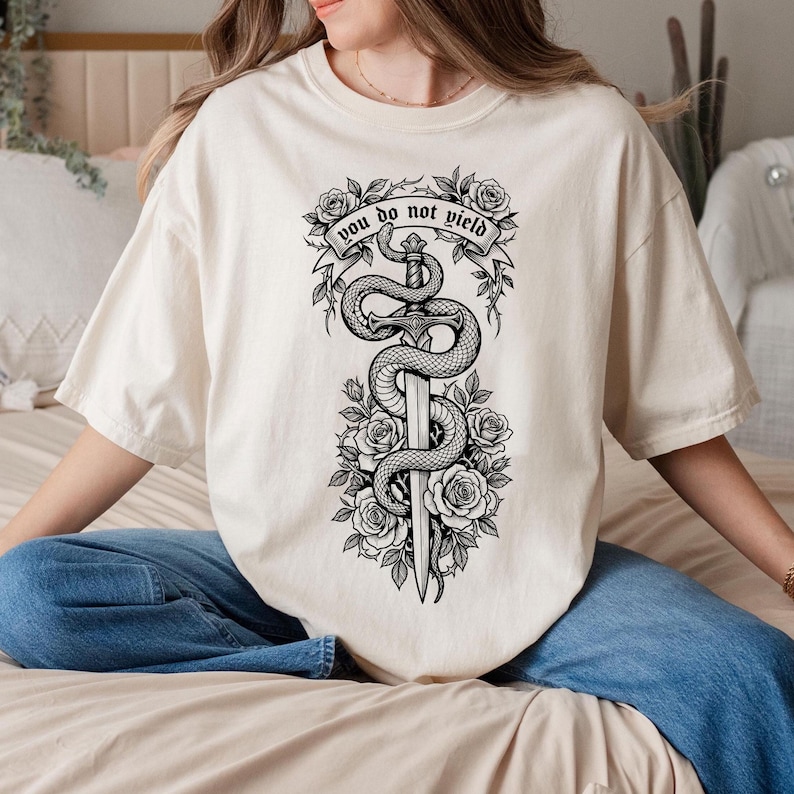 May include: Cream-colored t-shirt featuring a black and white graphic of a sword entwined by a snake, surrounded by roses. The text "you do not yield" is displayed on a banner above the sword. The shirt has short sleeves and a relaxed fit.