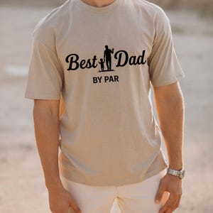 May include: A beige t-shirt with the words "Best Dad" in a cursive font, accompanied by a silhouette of a parent and child. Below the text is "BY PAR" in a sans-serif font. The shirt is worn by a person.