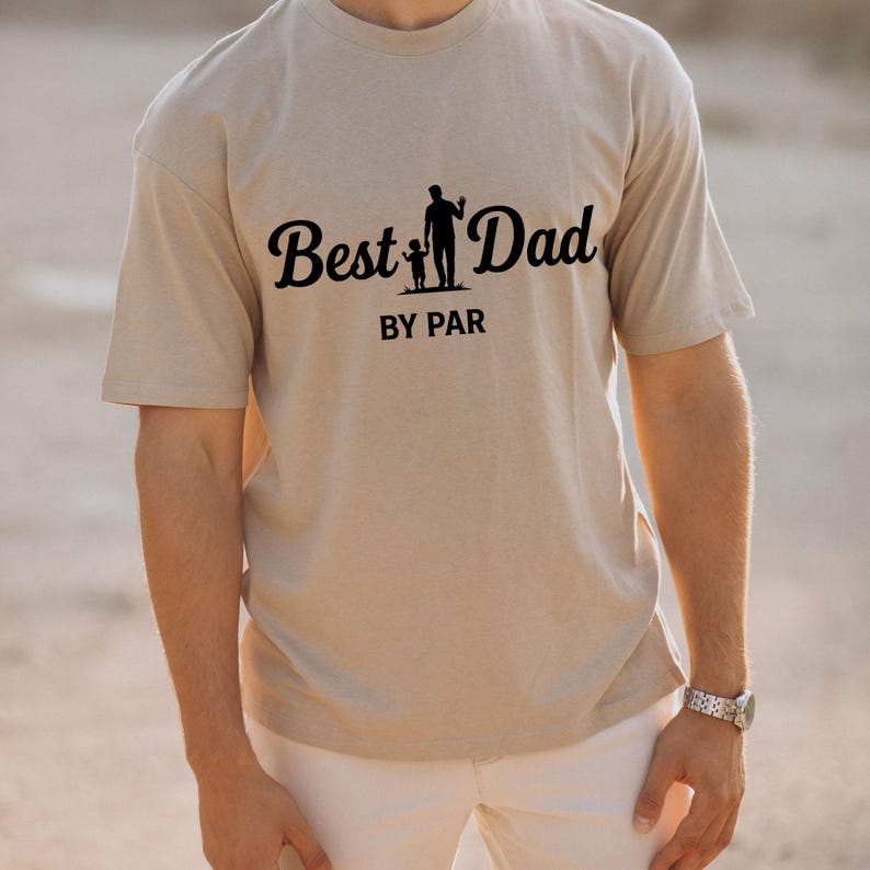 May include: A beige t-shirt with the words "Best Dad" in a cursive font, accompanied by a silhouette of a parent and child. Below the text is "BY PAR" in a sans-serif font. The shirt is worn by a person.
