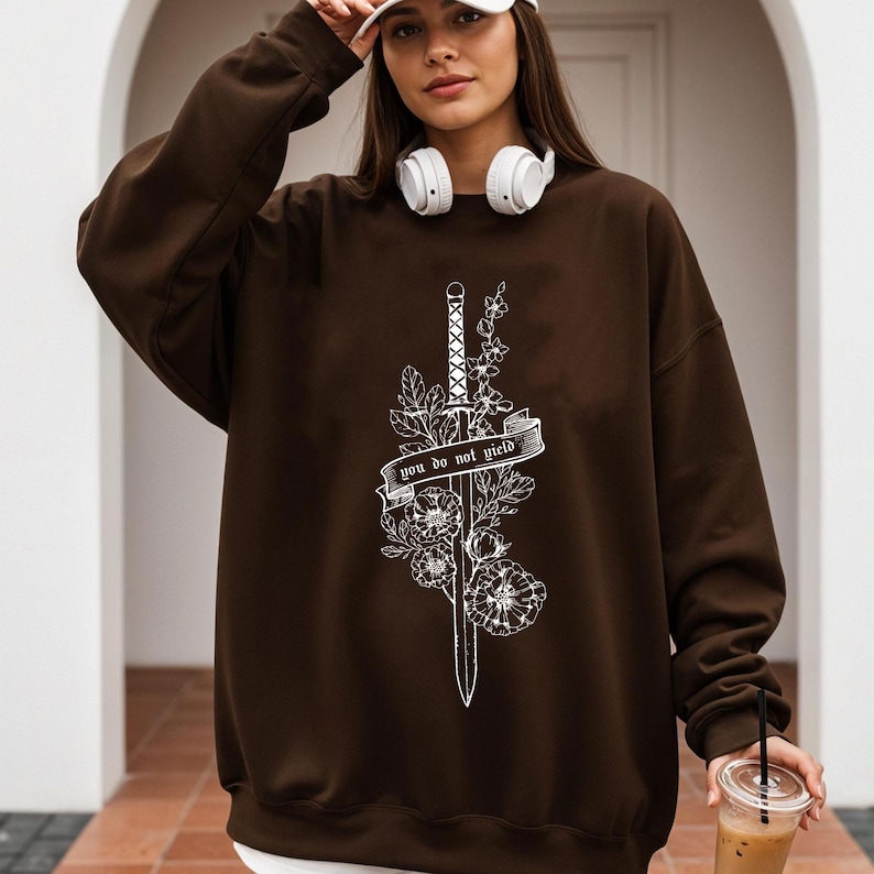 May include: A brown sweatshirt featuring a white floral and sword design with the text "you do not yield". The sweatshirt has long sleeves and a crew neck. The image shows a person wearing the sweatshirt.