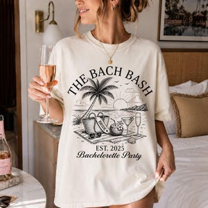 May include: An off-white t-shirt featuring a black graphic of a beach scene with a palm tree, a tote bag labeled BRIDE, a coconut drink, and glasses. The text reads THE BACH BASH, EST. 2025, and Bachelorette Party in a black serif and script font.