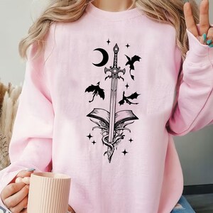 Puede incluir: Sudadera rosa claro con un dise&ntilde;o negro de una espada que atraviesa un libro abierto, con una luna creciente, estrellas y dragones. El dise&ntilde;o est&aacute; impreso en la parte delantera.