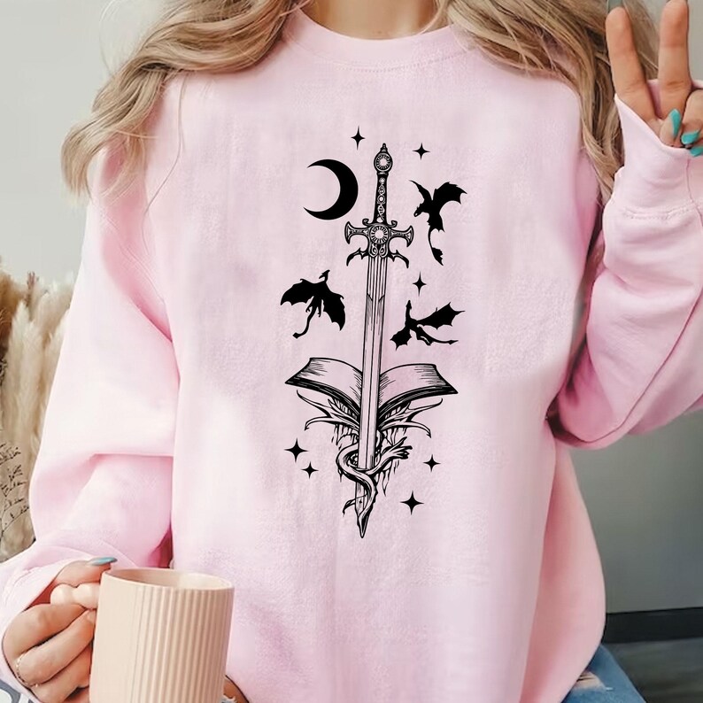 Puede incluir: Sudadera rosa claro con un dise&ntilde;o negro de una espada que atraviesa un libro abierto, con una luna creciente, estrellas y dragones. El dise&ntilde;o est&aacute; impreso en la parte delantera.
